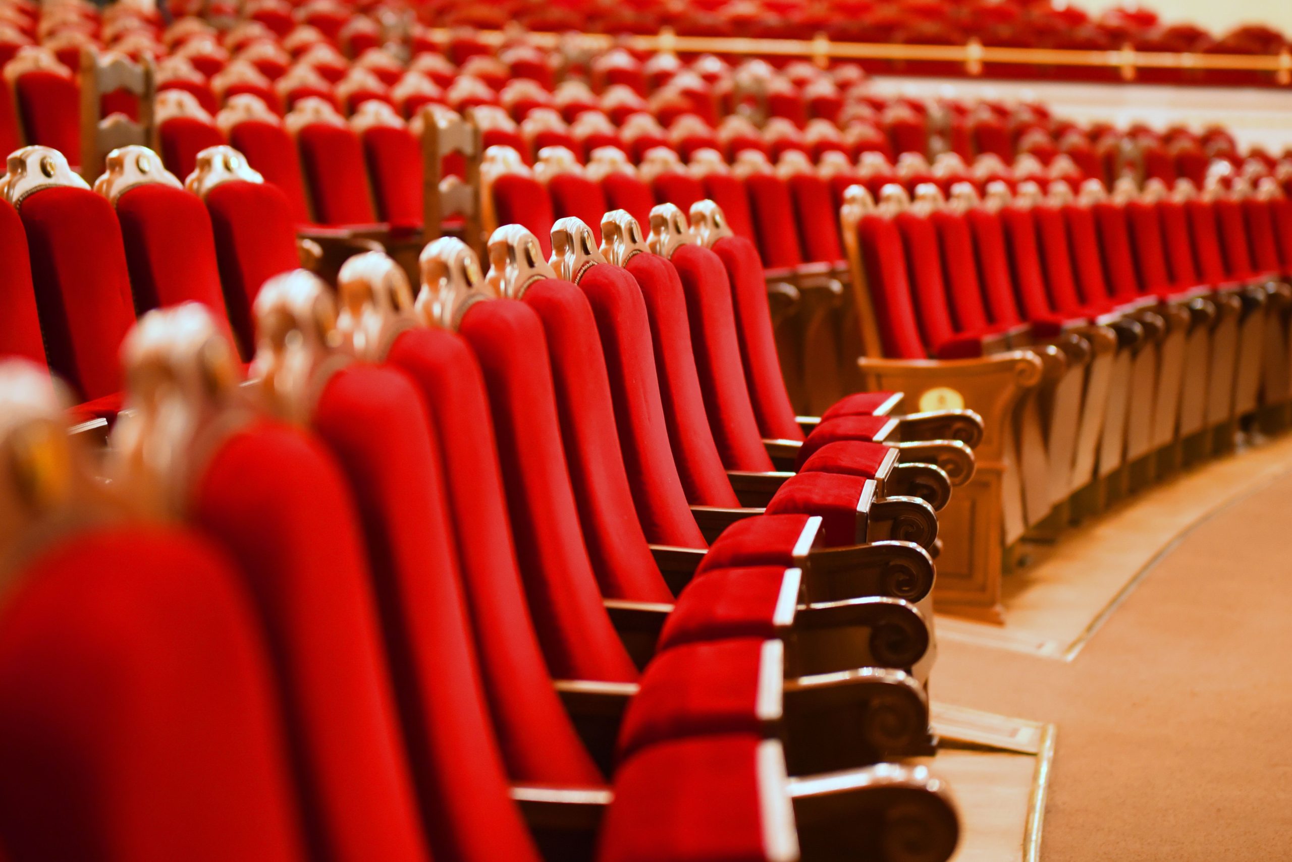 A photo of the interior of an empty theatre. The photo is cropped in tight on a row of empty red chairs stretch out into the dress circle