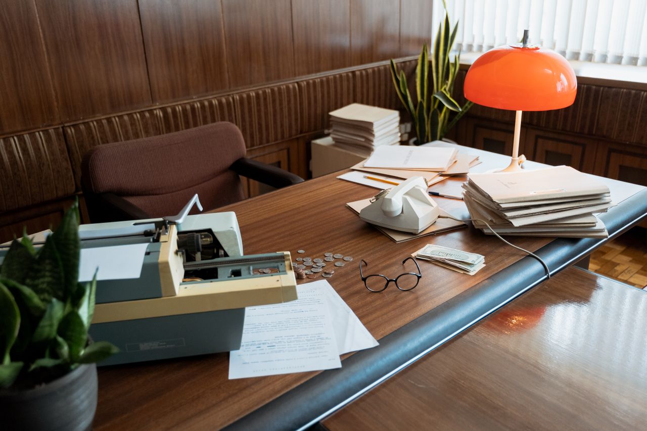 A photo of a desk with an electric keyboard, analogue phone, papers, a pair of glasses and an orange desk lamp strewn across it. The office and its furniture gives off a distinctly 1970s vibe